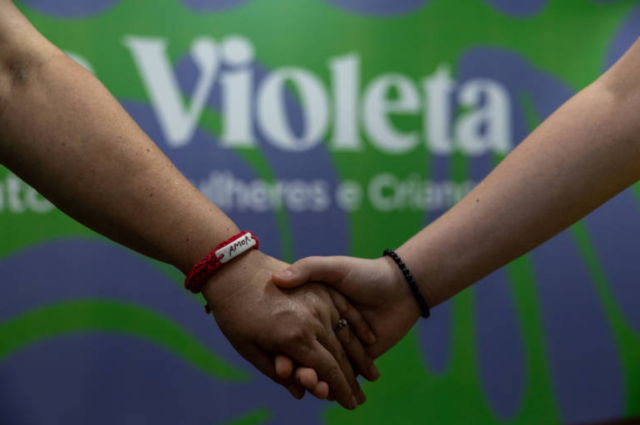 A imagem mostra duas mãos se segurando, com um fundo verde onde está escrito ‘Violeta’ em letras brancas. As mãos são de diferentes tonalidades de pele e estão unidas em um gesto de apoio ou amizade.